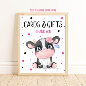 May include: A white sign with black polka dots and pink dots. The sign says "Cards & Gifts. Thank You" and features a cartoon black and white cow with a pink flower crown and a blue butterfly.