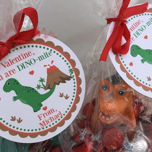 Puede incluir: Una bolsa de regalo para el D&iacute;a de San Valent&iacute;n con una cinta roja y una etiqueta redonda que dice "Valentine, you are DINO-mite" con un dinosaurio verde y un volc&aacute;n marr&oacute;n. La bolsa est&aacute; llena de caramelos rojos y plateados.