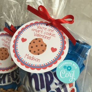 May include: A red and white Valentine's Day gift bag with a personalised tag that says "You're one smart cookie, Valentine!" with the name "Madison" printed below. The bag contains a bag of Chips Ahoy! cookies. The tag is tied with a red ribbon. A blue and white sticker with the text "PERSONALIZE WITH Coryl" is on the bag.