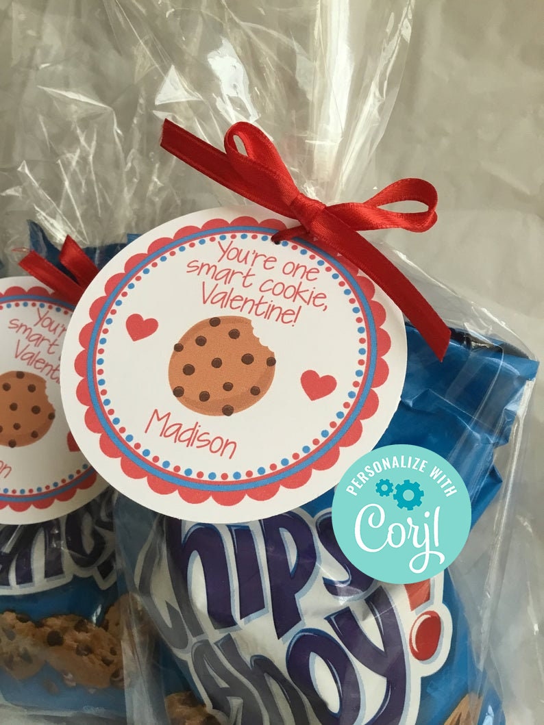 May include: A red and white Valentine's Day gift bag with a personalised tag that says "You're one smart cookie, Valentine!" with the name "Madison" printed below. The bag contains a bag of Chips Ahoy! cookies. The tag is tied with a red ribbon. A blue and white sticker with the text "PERSONALIZE WITH Coryl" is on the bag.