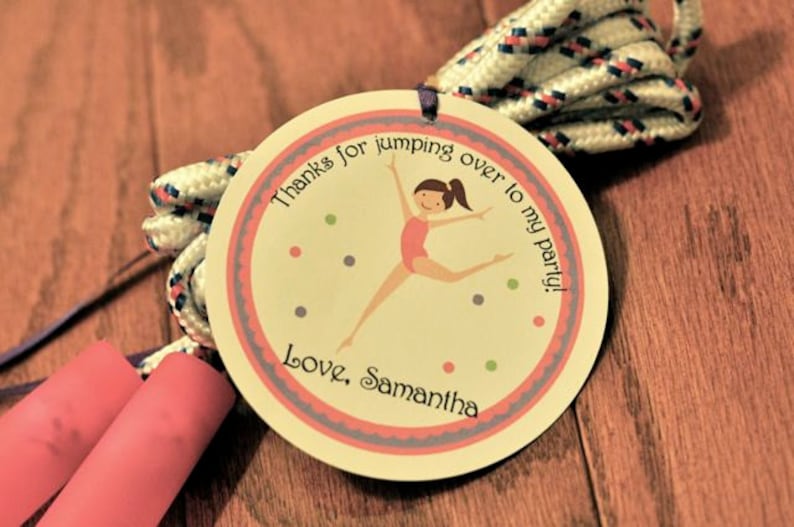 K&ouml;nnte beinhalten: Ein runder, cremefarbener Anh&auml;nger mit rosa Rand und lila Design. Er zeigt ein Cartoon-M&auml;dchen, das Seil springt, mit dem Text "Danke, dass du zu meiner Party gesprungen bist! Liebe, Samantha".