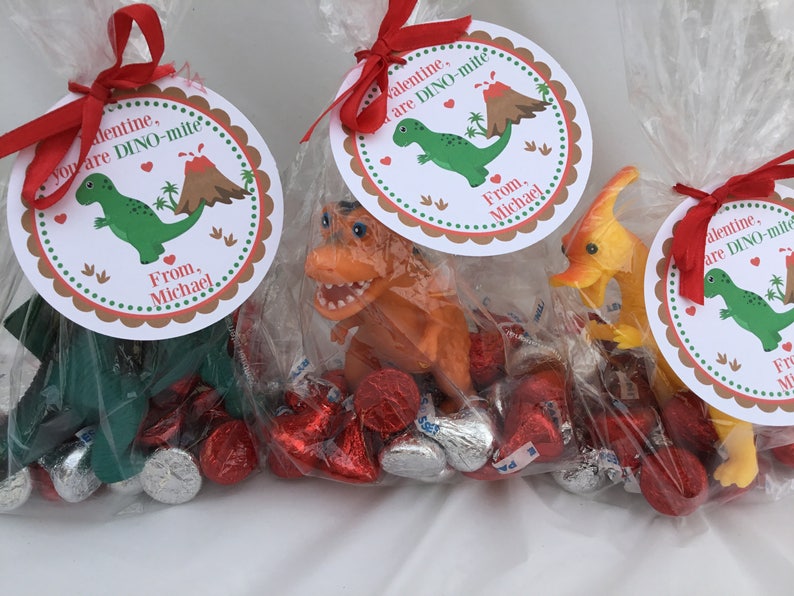 Puede incluir: Tres bolsas de pl&aacute;stico transparentes llenas de chocolates envueltos en papel de aluminio rojo y plateado. Cada bolsa tiene una etiqueta redonda de papel blanco con un dise&ntilde;o de dinosaurio y volc&aacute;n. Las etiquetas dicen "You are DINO-mite" y "From, Michael".