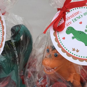 Puede incluir: Una bolsa de regalo para el D&iacute;a de San Valent&iacute;n con una cinta roja y una etiqueta que dice "Valentine, are DINO-mite!" La bolsa contiene un juguete de dinosaurio verde, un juguete de dinosaurio marr&oacute;n y corazones de caramelo rojos.