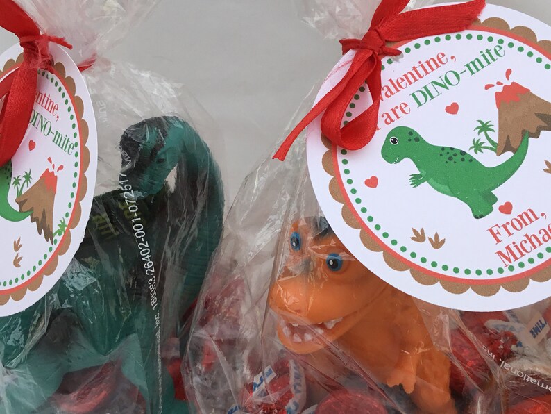 Puede incluir: Una bolsa de regalo para el D&iacute;a de San Valent&iacute;n con una cinta roja y una etiqueta que dice "Valentine, are DINO-mite!" La bolsa contiene un juguete de dinosaurio verde, un juguete de dinosaurio marr&oacute;n y corazones de caramelo rojos.