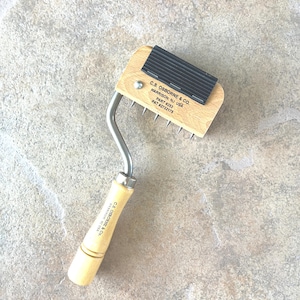 May include: A leatherworking tool with a wooden handle and a metal head. The head has a row of metal tines and a black rubber grip. The handle is light-colored wood with the text "C.S. OSBORNE & CO. HARRISON, NJ USA PART #253 PAT #2732175".