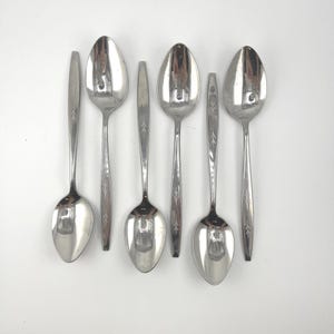 May include: Six stainless steel spoons arranged on a white surface. The spoons have a reflective, polished finish and a simple, elegant design. Each spoon features a decorative etched pattern on the handle.