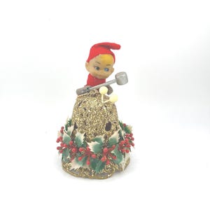 May include: A vintage Christmas bell ornament featuring a small elf figure in a red hat and shirt, holding a silver hammer. The bell is gold with glitter, holly, and red berry accents. A festive holiday decoration.