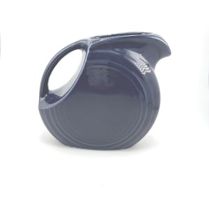 May include: A navy blue ceramic pitcher with a large handle and a spout. The pitcher has a rounded body with concentric rings near the base. The handle is a large, open loop. The pitcher is on a white background.