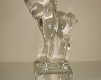 May include: A clear glass figurine of a young horse standing on a rectangular base. The sculpture features a smooth, transparent surface with defined contours representing the mane, ears, and tail of the foal, capturing light and creating shadows.