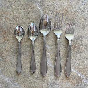 May include: Five pieces of stainless steel silverware, including three spoons and two forks, are arranged on a textured, neutral-toned surface. The spoons vary in size, and the forks have four tines each. The silverware reflects the surrounding environment.