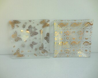 May include: Two square glass plates with gold designs. One plate features gold butterflies, suns, and leaves. The other plate has a gold geometric pattern. The plates are likely decorative or for serving.