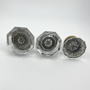 May include: Three clear glass doorknobs with octagonal and round shapes. The doorknobs have a decorative, starburst-like design in the center and are set against a white background.