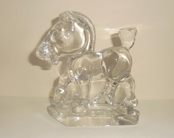 May include: A clear glass horse figurine. The horse is depicted in a seated position with its head turned to the left. The figurine has a detailed mane and tail, and sits on a rectangular base. The glass is transparent.