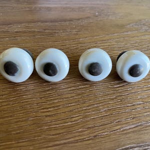 May include: Four round cabinet knobs featuring a smooth white ceramic surface with a dark metallic center button. Each knob has a circular profile and a polished finish, designed for attachment to furniture doors or drawers for functional use.