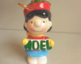 May include: A ceramic Christmas ornament featuring a cartoon character wearing a red hat and a red and yellow shirt. The ornament holds a sign that reads "NOEL" in green letters. The character has black hair and a gold string for hanging.