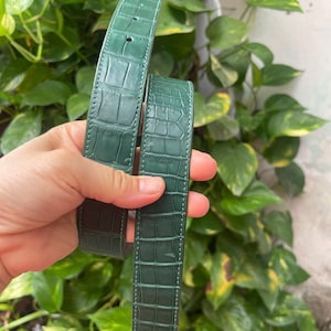 Puede incluir: Un cinturón de color verde oscuro con un patrón texturizado de piel de cocodrilo. El cinturón tiene costuras visibles a lo largo de los bordes y una serie de agujeros preperforados para el ajuste. La superficie tiene un acabado brillante.