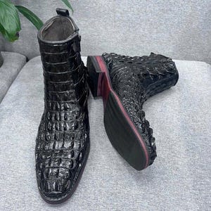 Könnte beinhalten: Ein Paar schwarze Stiefeletten mit einem glänzenden, strukturierten Krokodilleder-Muster. Die Stiefel verfügen über einen seitlichen Reißverschluss, eine abgerundete Zehenpartie und einen niedrigen Absatz mit einer roten Akzentlinie an der Sohlenkante.