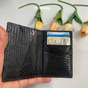 Pode incluir: Carteira de couro preto texturizado, aberta para exibir ranhuras para cartões e um bolso. A carteira tem um padrão semelhante ao de um réptil. Cartões com texto são visíveis nas ranhuras. As tulipas artificiais estão no fundo.