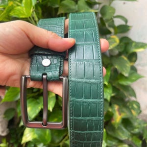 Puede incluir: Un cinturón verde bosque con un patrón texturizado de piel de cocodrilo. El cinturón incluye una hebilla rectangular de metal gris oscuro mate y un remache metálico plateado que asegura la correa cerca de la hebilla.