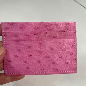 May include: A pink card holder with a textured ostrich leather design. The card holder has three card slots and is stitched with white thread. The pink leather has a pattern of small, raised bumps.