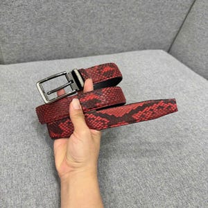 Pode incluir: Um cinto vermelho e preto com padrão de pele de cobra e fivela retangular prateada. O cinto é feito de um material texturizado. Acessório de moda, em fundo cinza.