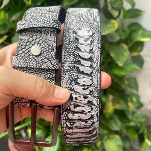 Op de afbeelding: Een riem met een gestructureerd zwart-wit patroon dat lijkt op struisvogelhuid. De band is voorzien van een zilverkleurige metalen stud op de lus en een donkere, matte metalen gesp. Het oppervlak heeft een duidelijk verhoogde textuur.