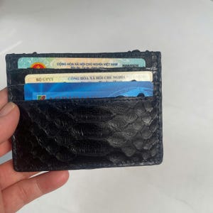 May include: A black, textured leather cardholder with multiple slots, holding several cards. The cardholder has a scale-like pattern and is designed to carry credit cards, IDs, or business cards. The cardholder is held in a hand.