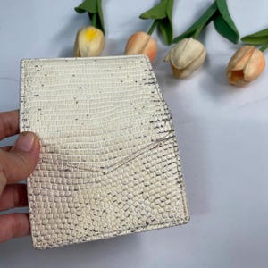 Pode incluir: Um porta-cartões branco com textura de pele de réptil. O porta-cartões é segurado por uma mão e tem um bolso na frente. O fundo inclui flores artificiais com caules verdes e pétalas amarelas, laranjas e brancas.