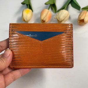 Può includere: Portacarte in pelle arancione strutturata con una carta blu parzialmente visibile. Il portacarte è rettangolare e ha un interno rosso. La parola "mbank" è visibile sulla carta.