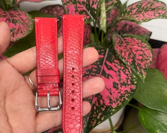 Puede incluir: Una correa de reloj roja de dos piezas con un patrón texturizado similar a escamas en la superficie. La correa incluye una hebilla de metal plateado, dos trabillas fijas y una serie de orificios perforados para el ajuste en color rojo.