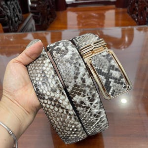 Puede incluir: Un cinturón con un patrón texturizado de piel de serpiente en tonos gris, negro y blanco. El cinturón incluye una hebilla rectangular de metal dorado con una incrustación de piel de serpiente a juego y un mecanismo deslizante.