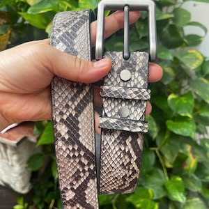 Puede incluir: Un cinturón con estampado de piel de serpiente en tonos beige, gris y negro. El cinturón incluye una hebilla rectangular de metal plateado cepillado y dos trabillas a juego, aseguradas con dos remaches de metal plateado cerca de la hebilla.