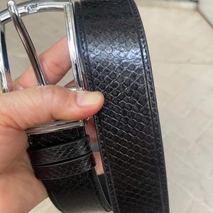 Puede incluir: Un cinturón negro con un patrón texturizado de piel de serpiente. El cinturón tiene una hebilla de color plateado. El cinturón está en la mano de una persona.