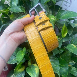 Puede incluir: Un cinturón de color amarillo brillante con un patrón texturizado de piel de cocodrilo. El cinturón incluye una hebilla rectangular de metal gris mate, trabillas amarillas a juego y costuras visibles a lo largo de los bordes de la correa.