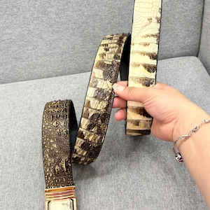 Op de afbeelding: Een riem met een gestructureerde, donkerbruine band met krokodillenpatroon die overgaat in een lichter, crèmekleurig gedeelte met grote, rechthoekige schubben. De riem is voorzien van een rechthoekige, goudkleurige metalen gesp met embleem.
