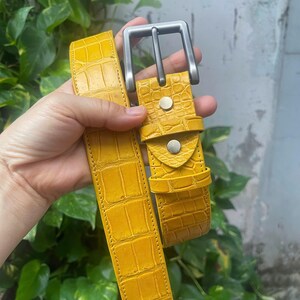 Op de afbeelding: Een felgele riem met een gestructureerd krokodil-reliëfpatroon. De riem is voorzien van een matzilverkleurige metalen gesp, twee bijpassende zilverkleurige metalen klinknagels bij de gesp en een bijpassende gele riemlus van kunstleer.
