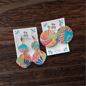 May include: Handmade earrings with colorful floral and geometric patterns. The earrings are displayed on white cards with the text "Tiendita hand made jewelry" and "Con Amor Isa Monique". The earrings have gold-colored hooks.