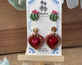 May include: A pair of handmade earrings featuring a red heart design with gold accents and a green cactus design with pink accents. The earrings are displayed on a white card with the text "Tiendita - hand-made jewelry" and "Con Amor Lisa Monique".
