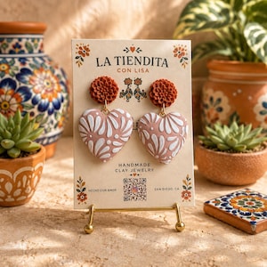 May include: A pair of heart-shaped clay earrings on a display card labeled LA TIENDITA CON LISA. The earrings feature a dusty pink heart with white floral patterns and a textured, rust-colored flower stud. The card includes a QR code and text.