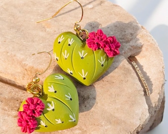 May include: A pair of heart-shaped earrings with a cactus design. The earrings are lime green with white accents and small pink flowers. The earrings have gold-colored hooks.