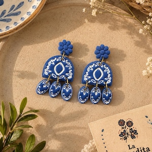 May include: A pair of blue and white dangle earrings featuring a flower-shaped stud, a semi-circular piece with intricate white patterns, and three small oval charms hanging from the bottom, all connected by small gold-toned metal jump rings.