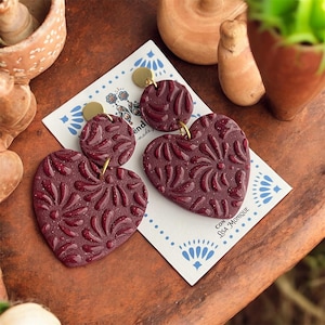 May include: Burgundy heart-shaped earrings with a floral pattern and gold-colored accents. The earrings are on a white card with blue floral designs and the text "Lisa Monique". The earrings are displayed on a brown surface.