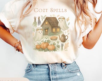 May include: Ivory-colored t-shirt with a whimsical illustration featuring a cozy cottage, pumpkins, ghosts, and the text "Cozy Spells." The design includes a teapot, pie, and floral accents, perfect for autumn.