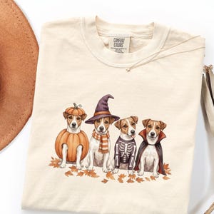 May include: Cream-colored t-shirt featuring a Halloween-themed graphic. The graphic depicts four dogs dressed in Halloween costumes: a pumpkin, a witch, a skeleton, and a vampire. The shirt has a Comfort Colors tag.