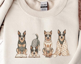 May include: A cream-colored sweatshirt featuring a watercolor illustration of four Australian Cattle Dogs. Each dog is depicted in a different pose, with one reading a book, and another wrapped in a blanket. The sweatshirt has a crew neck and long sleeves.