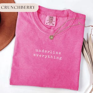 May include: A bright pink t-shirt with the words "underline everything" in white lettering. A gold necklace with a pendant rests on the shirt. The shirt is folded and has a tag that reads "Comfort Colors".