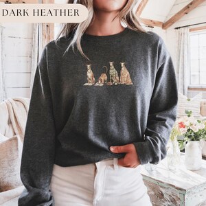 May include: Dark heather gray sweatshirt featuring an embroidered design of four dogs in various poses. One dog is wearing a plaid sweater, and another is draped in a blanket. The text "DARK HEATHER" is in the upper left corner.