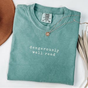 May include: A seafoam green Comfort Colors t-shirt with the words "dangerously well read" in white. A silver necklace with a small pendant rests on the shirt. The shirt is folded neatly.