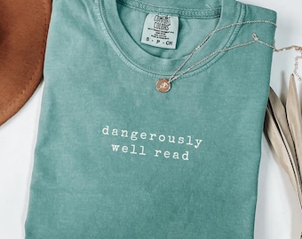 May include: A seafoam green Comfort Colors t-shirt with the words "dangerously well read" in white. A silver necklace with a small pendant rests on the shirt. The shirt is folded neatly.