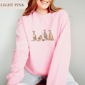 May include: Light pink sweatshirt with a crew neck. The sweatshirt features an illustration of four greyhounds in various poses. The text "LIGHT PINK" is in a white rectangle in the upper left corner.